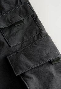 Black fabric with a smooth texture, featuring two flap pockets, and contrasting stitching on the sleeve. Minimalist design, functional details.