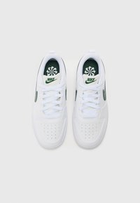 Baskets blanches avec une tige en cuir lisse, des accents verts et des lacets texturés. Elles présentent des perforations sur le bout et une languette de marque.