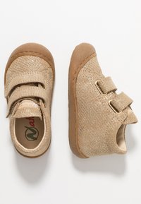 Beige Kinderschuhe mit strukturierter Oberfläche, ausgestattet mit zwei Klettverschlüssen und einer Gummisohle. Runde Zehenform und niedrig geschnittenes Design für Komfort.