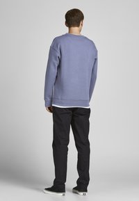 Felpa viola con colletto a girocollo, polsini e orlo a coste, indossata sopra una camicia bianca, abbinata a pantaloni neri e sneaker nere.