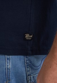 Mornarsko modra bombažna majica s majhno črno oznako z napisom "Petrol Industries" v beli barvi. V kombinaciji z zbledelimi modrimi denim kratkimi hlačami.