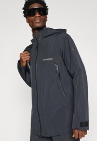 Muž v čiernej bundě Peak Performance s kapucňou, ktorý drží popruh batohu a má na sebe veľké čierne slnečné okuliare na jednoliatom pozadí.