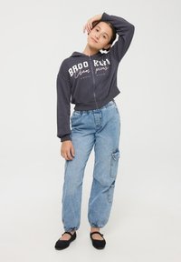 Przycięta ciemnoszara bluza z kapturem na suwak z napisem "BROOKLYN", połączona z jasnoniebieskimi spodniami cargo i czarnymi balerinami. Luźny krój, elastyczne mankiety.