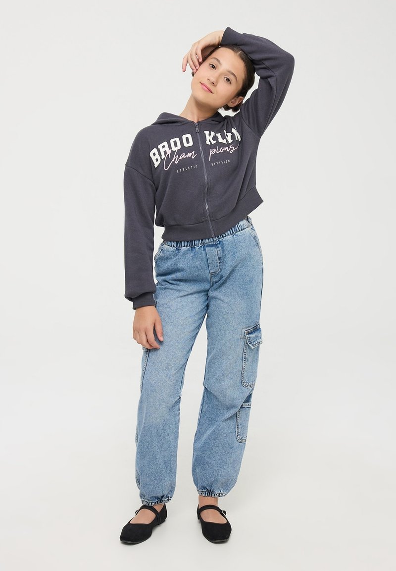 Przycięta ciemnoszara bluza z kapturem na suwak z napisem "BROOKLYN", połączona z jasnoniebieskimi spodniami cargo i czarnymi balerinami. Luźny krój, elastyczne mankiety.