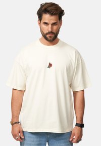 Cremefarbene BaumwollT-Shirt mit kurzen Ärmeln, das ein kleines gesticktes Design auf der Vorderseite hat. Lockere Passform und Rundhalsausschnitt.