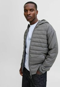 Grå pufferjakke med vandret quiltet design, lynlåslukning foran, fast hætte, parret med en lys grå T-shirt og mørke jeans.