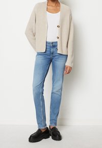 Cardigan beige lavorato a maglia con tre bottoni, indossato sopra una maglietta bianca e jeans azzurrini. Mocassini neri con tacco alto completano l'outfit.