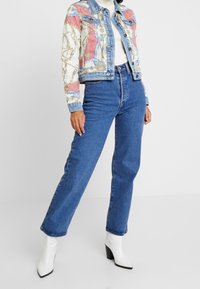 Veste en denim avec un imprimé chaîne coloré et un corps bleu, associée à un jean bleu taille haute et des bottines à talons blancs.
