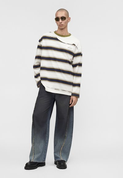 Άνδρας με υπερμεγέθη λευκό πουλόβερ με ναυτικού και χρυσού οριζόντιες ρίγες, φαρδιά φθαρμένα μαύρα τζιν, μαύρα παπούτσια και ορθογώνια γυαλιά ηλίου.