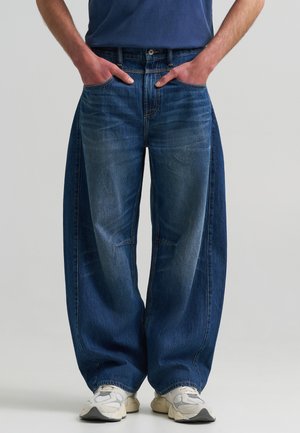 Homme portant un jean large bleu foncé, les mains dans les poches et des baskets blanches, debout sur un fond uni gris clair.