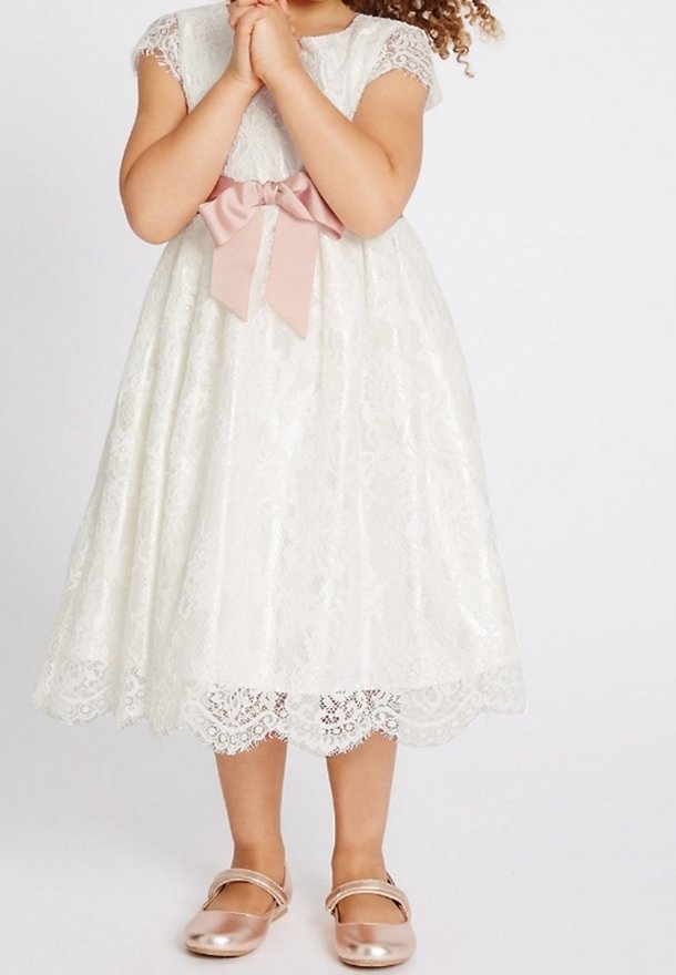 Fille portant une robe en dentelle blanche avec un nœud rose à la taille et des chaussures Mary Jane roses métalliques, mains jointes près de la poitrine.