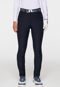 Slim-fit marinblå byxor med ett vävt bälte, tillsammans med vita och blå sneakers, en vit långärmad topp och en vit golfhandske på höger hand.