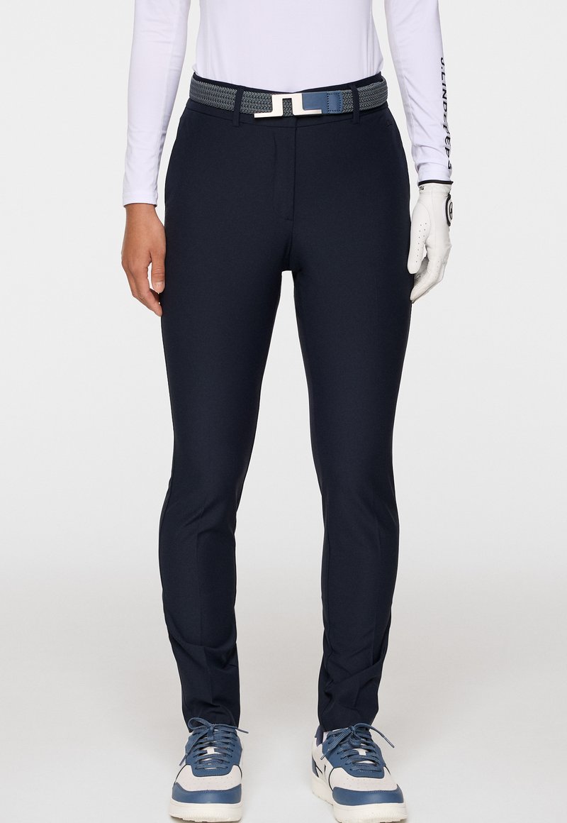 Slim-fit marinblå byxor med ett vävt bälte, tillsammans med vita och blå sneakers, en vit långärmad topp och en vit golfhandske på höger hand.