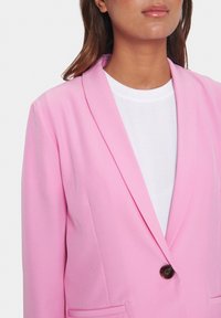 Růžový blazer s šálovým límcem, jedním hnědým knoflíkem a klopovými kapsami. Nosený přes bílý tričko, vyrobené z hladkého materiálu.