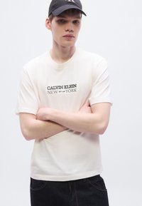 Μπεζ βαμβακερό μπλουζάκι με κοντά μανίκια, με μαύρο τυπωμένο λογότυπο "Calvin Klein" και "NEW YORK" σε κεντρικό σχέδιο.