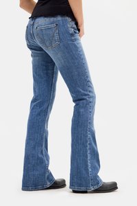 Osoba v modrých rozšírených džínsoch a čiernych čižmách, stojaca so založenými rukami na páse na jednolatom pozadí.