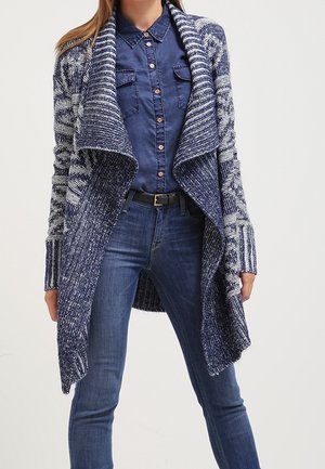 Femme portant un cardigan bleu à motifs ouvert sur le devant, une chemise bleu foncé boutonnée et un jean bleu avec une ceinture noire.