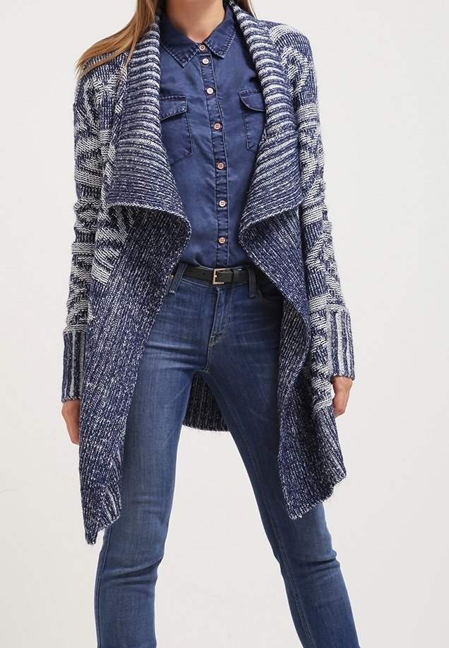 Femme portant un cardigan bleu à motifs ouvert sur le devant, une chemise bleu foncé boutonnée et un jean bleu avec une ceinture noire.