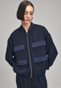 Giacca a bomber blu navy con zip frontale, due grandi tasche blu, polsini a costine e un tessuto leggero. Design semplice, vestibilità comoda.