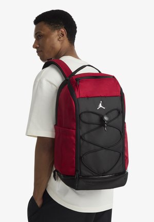 Sac à dos rouge et noir en tissu texturé, avec des bretelles réglables, une poche zippée à l'avant et des détails élastiques à cordon bungee.