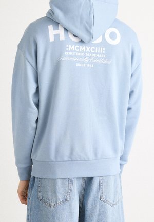Person trägt eine hellblaue Kapuzenjacke mit weißem Text "HUGO MCMXCIII Registered Trademark Since 1993" auf der Rückseite, kombiniert mit hellblauen Jeans.