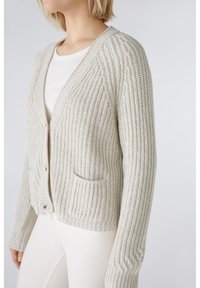 Hellbeige gestrickte Strickjacke mit einem strukturierten Muster, V-Ausschnitt, zwei vorderen Taschen und Knopfverschluss. Die langen Ärmel verfügen über gerippte Bündchen.