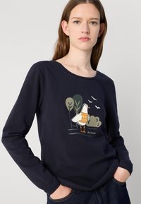 Tumesinise pika varrukaga särk, millel on linnu graafika maa toonides. Pehme kangas, ümmargune kaelus ja mugav lõige. Brändi logo on nähtav.