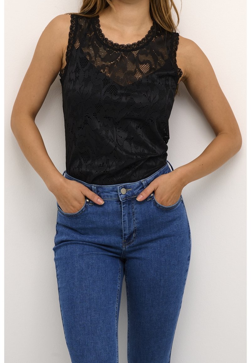 Musta pitsinen tankkitoppi, jossa on kukkakuosi ja hapsureunat, yhdistettynä korkeavyötäröisiin sinisiin farkkuihin, joissa on etutaskut ja napitus.