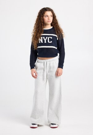 Jeune personne aux longs cheveux bouclés portant un sweat-shirt bleu marine "NYC", un pantalon de jogging à jambes larges gris clair et des baskets noires et blanches, debout et regardant vers la droite.