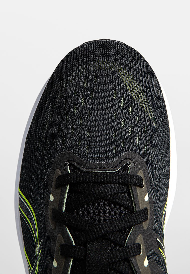 Zapatilla deportiva negra con parte superior texturizada, que presenta secciones de malla para la transpirabilidad, detalles en verde y una gruesa suela blanca.