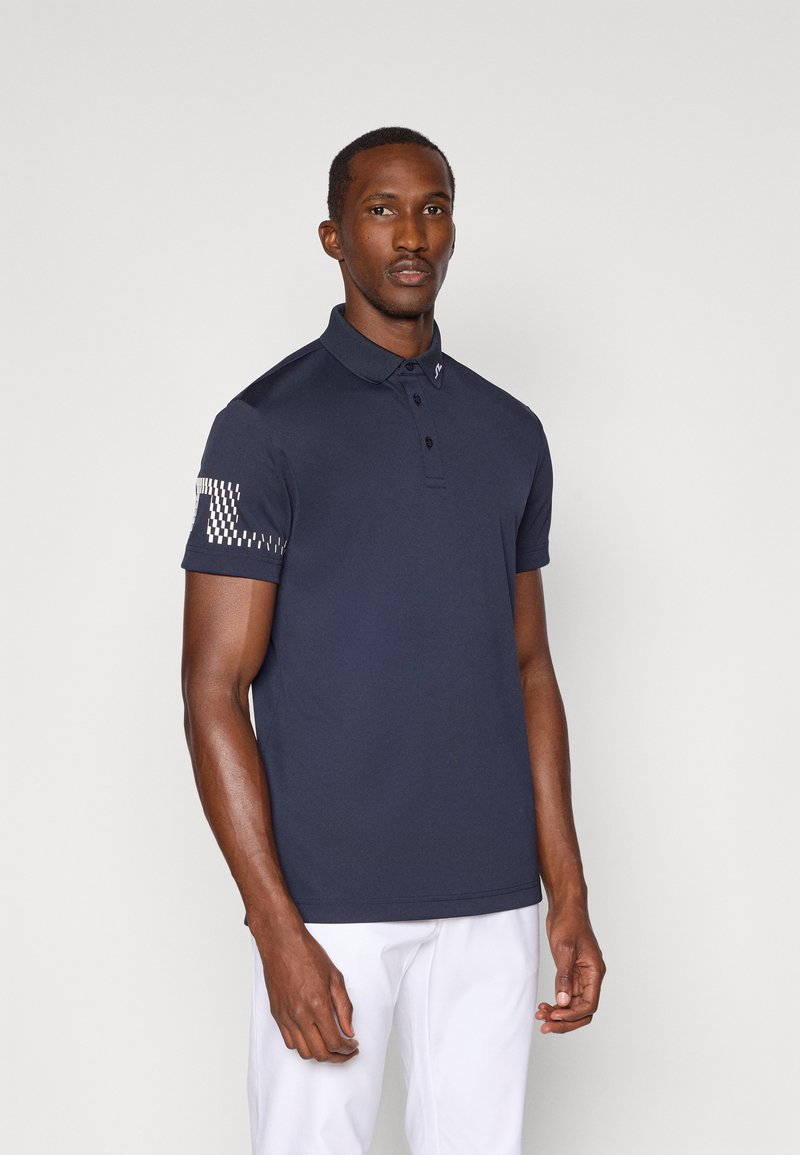 Marineblå polo-skjorte laget av glatt materiale, med en to-knapps knappestolpe og et teksturert mønster på venstre erme.