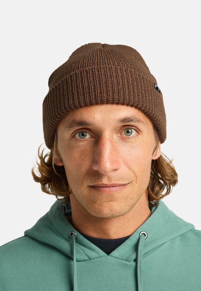 Gorro de punto marrón con textura acanalada y un pequeño parche de logo, llevado por una persona con una sudadera color turquesa, con un fondo neutro.