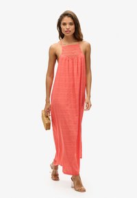 Abito maxi corallo con un corpetto testurizzato, spalline sottili e una silhouette fluida; abbinato a sandali con tacco a blocco trasparenti e una borsa intrecciata.