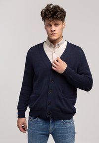 Granatowy kardigan z głębokim dekoltem w kształcie litery V, z przodu zapinany na guziki i dwoma kieszeniami. Dzianinowa faktura oraz ściągacze przy mankietach i dole. Nosi się go na jasnej koszuli.