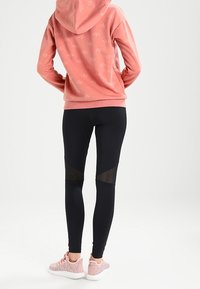 Rosa hoodie med huva, mönstrad med en diskret logotyp, tillsammans med svarta leggings med meshdetaljer och lätta rosa skor.