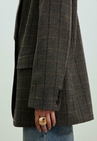 Blazer à motif pied-de-poule en marron et gris, avec une coupe ajustée, des poignets boutonnés et une poche poitrine. Bague en or au doigt.