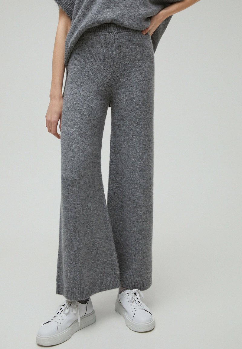 Femme portant un pantalon en maille gris ample et des baskets blanches à plateforme, debout avec une main sur la hanche sur un fond uni.