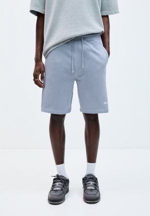 Pantaloni corti in cotone azzurro chiaro con vita elasticizzata e lacci, dotati di tasche laterali. Abbinati a sneakers scure e calze bianche alla caviglia.