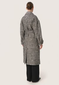 Lang, houndstooth-gepatenteerde jas in zwart en wit, met een brede kraag, ceintuur en oversized mouwen. G gedragen over brede zwarte broek.