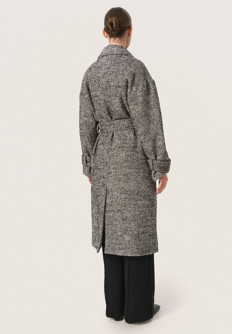 Abrigo largo de patrón de espiga en blanco y negro, con un cuello amplio, cinturón y mangas oversize. Llevado sobre pantalones negros de pierna ancha.