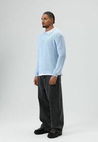 Jonge man die staat, gekleed in een lichtblauwe gebreide trui, loszittende zwarte jeans, zwarte schoenen, met cornrow-vlechtjes tegen een effen grijze achtergrond.