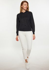 Fekete pamut kapucnis pulcsi kötéllel, fehér slim-fit nadrággal és fehér sportcipővel párosítva, egy sima háttér előtt állva.