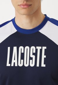 Temno modra majica z belimi detajli, s poudarjenim napisom "LACOSTE". Material je teksturiran, z okroglim ovratnikom in kontrastnimi rokavi.