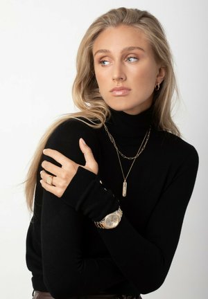 Sort turtleneck-sweater, lange ærmer, guldkæder, guldur med sølvdetaljer, ring på hånden, lys baggrund.