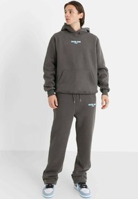 Sudadera gris con joggers a juego, fabricados en un material suave. Presenta un bolsillo frontal, logo en azul claro y un corte relajado. Zapatillas con acentos azules.