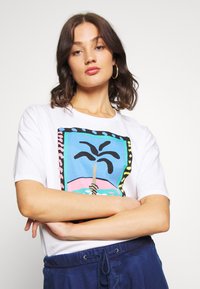 Camisa de tirantes blanca con un gráfico colorido de una palmera dentro de un marco rectangular y acentos con patrones. Material de algodón suave.