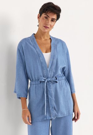 Femme portant une veste en denim bleu clair à manches 3/4 nouée à la taille, assortie à un pantalon en denim et un haut blanc en dessous.