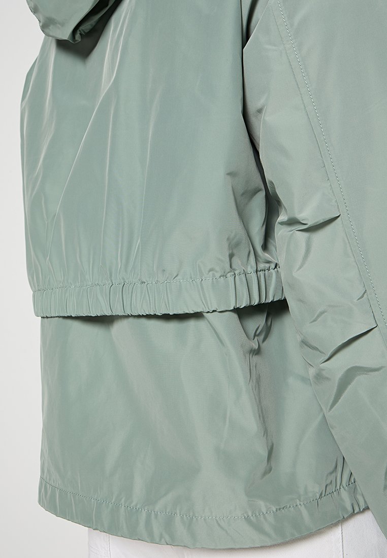 Hellgrüne, wasserabweisende Jacke mit elastischem Bund, Seitentaschen und glatter, glänzender Textur. Verstellbare Kapuze enthalten.