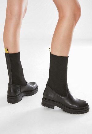 Botas de cuero negro hasta el tobillo con un eje de punto negro acanalado, que cuentan con una tira amarilla en el talón y una suela de goma negra texturizada con detalles de tracción.