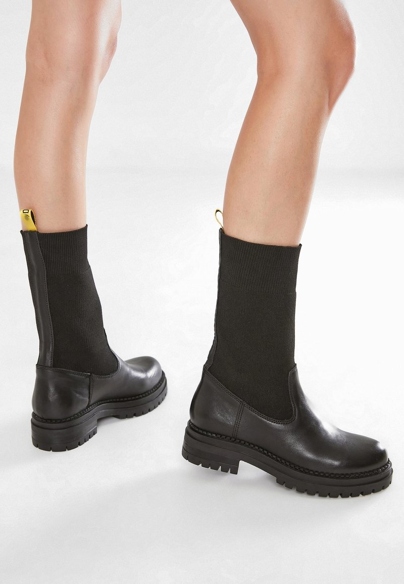 Svarta läderankelboots med skaft i ribbat svart stickat material, utrustade med en gul dragtab och en texturerad svart gummisula med mönsterdetaljer.